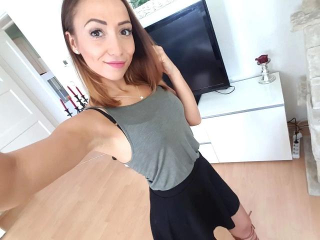 LeeTizia, aantrekkelijk met een gemiddelde borstmaat, draagt een grijze top en een zwarte rok, maakt een selfie in een lichte kamer, glimlacht met roze lippenstift, heeft bruin haar en een witte tv-kast..