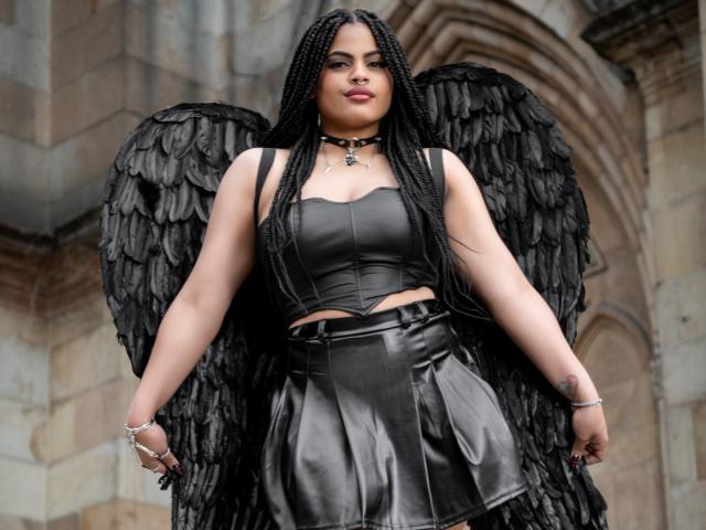 AprilKershaw with black wings, long braided hair, black leather crop top, black leather skirt, choker, and bracelets, standing confidently, showing off her tits and black feathered wings.