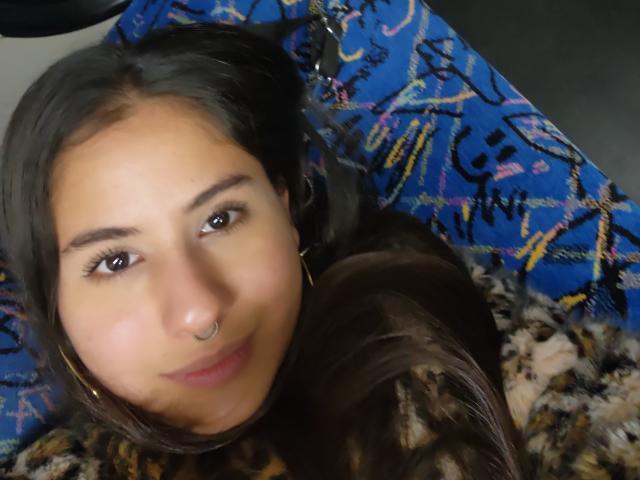 AuroraMonroe has tan skin, dark brown eyes, and a nose ring. She's smiling with long brown hair, wearing a leopard print shirt, and lying on a blue patterned blanket.