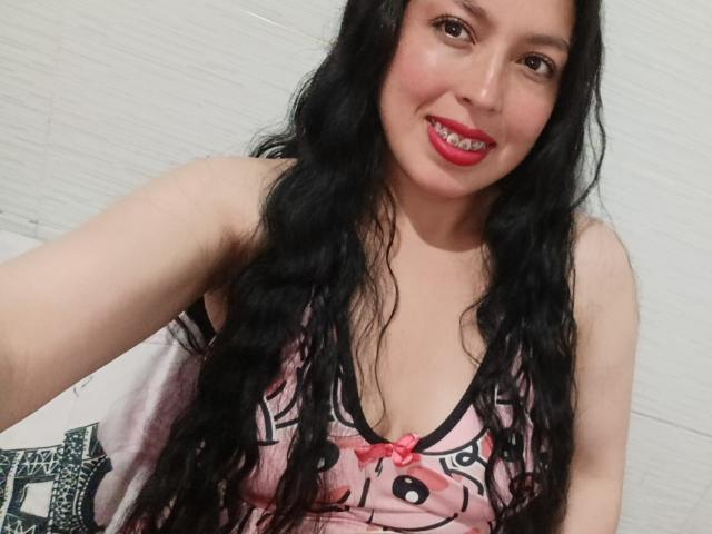 SylvieColette, con cabello negro largo y ondulado, lápiz labial rojo, sonriendo, vestida con una blusa rosa estampada con detalles negros, que deja ver sus pequeños senos, sosteniendo una cámara para tomarse un selfie..