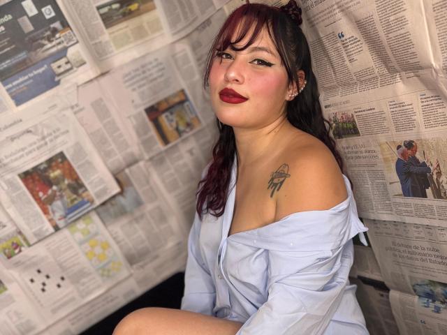 DominicTheOne, with dark red lipstick and black eyeliner, sits wearing an off-shoulder blue shirt, exposing a tattoo on her left shoulder, and has purple-dyed hair.