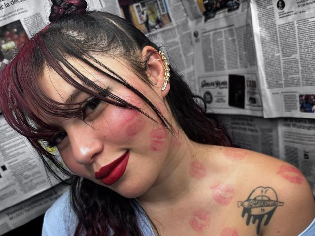 DominicTheOne with red lipstick, black and red hair, ear studs, red kiss marks on face and chest, black tattoo on shoulder, smiling at camera, newspaper in background.