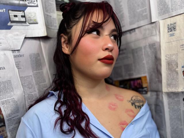 DominicTheOne, with dark red hair in a ponytail, has pink lipstick kisses on her chest, wearing a blue shirt, and has a tattoo of a small animal on her right shoulder.