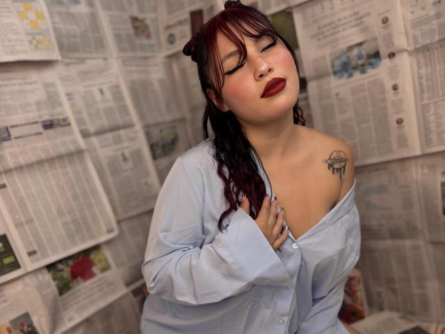DominicTheOne with dark red hair, eyes closed, red lipstick, light blue shirt, exposing left shoulder tattoo, hand on chest, medium tits, soft light, relaxed expression, paper-covered background.