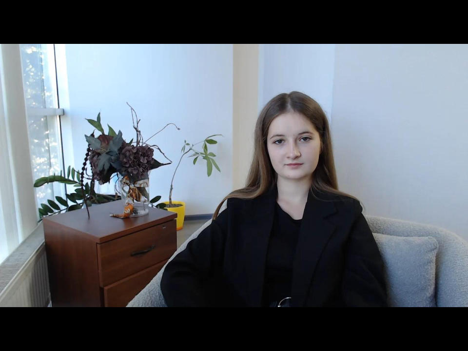MiyaLi has long brown hair, a black blouse, and a neutral expression, sitting in a gray chair, with a wooden dresser and plants to her left.
