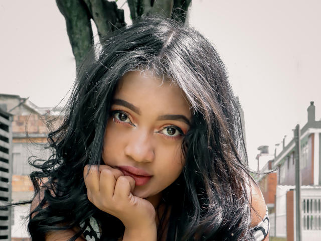 SaritaCute has long black wavy hair, brown eyes, and is resting her chin on her hand, looking up with a slightly pouty expression, showing her nose and lips.