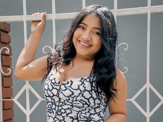 Sarita, carina, con capelli neri ondulati, sorridente, che tiene un vestito bianco e nero, mostrando il seno, in piedi vicino a una recinzione di metallo bianco e a un muro di mattoni..