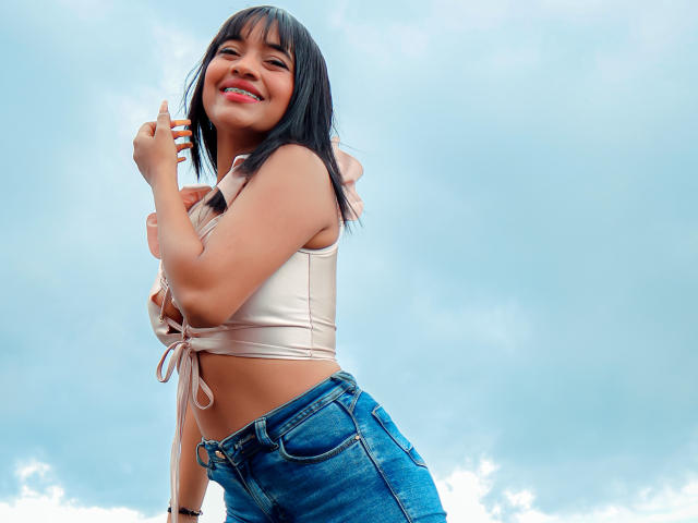 SaritaCute has medium tits, black hair, and a cute smile. She's wearing a tied-up white top and blue jeans, holding a striped drink with a cloudy sky.