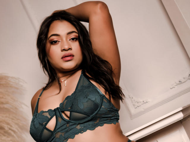 SaritaCute with dark hair, brown eyes, and full lips, wearing a green lace bra, has one arm raised, showing her tits, and a gold necklace.
