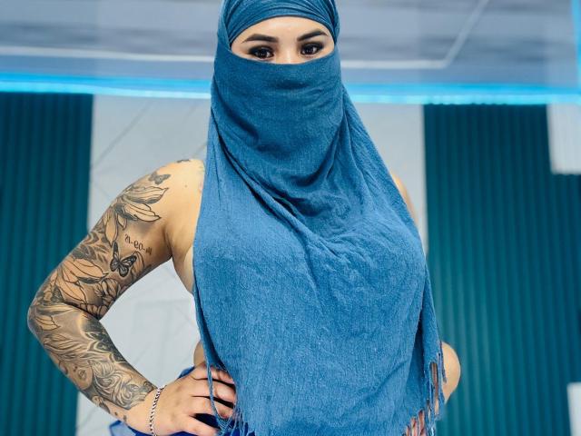 MalakCote with a blue scarf covering her face, showing tattooed arms and a hand on her hip. She has brown eyes and blue scarf. Her tits are visible.