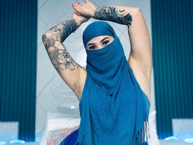 MalakCote with blue mask covering face, arms raised, showing tattooed sleeves, wearing matching blue scarf, exposing armpits, brown eyes, in a blue-themed setting.