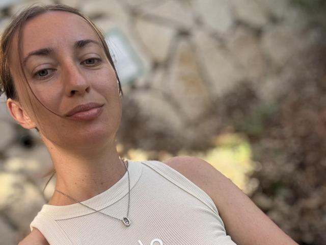 AskaSunshine with light brown skin and brown hair, wearing a sleeveless white top, necklace, slight smile, and subtle makeup, looking at the camera. Her small tits are barely visible.