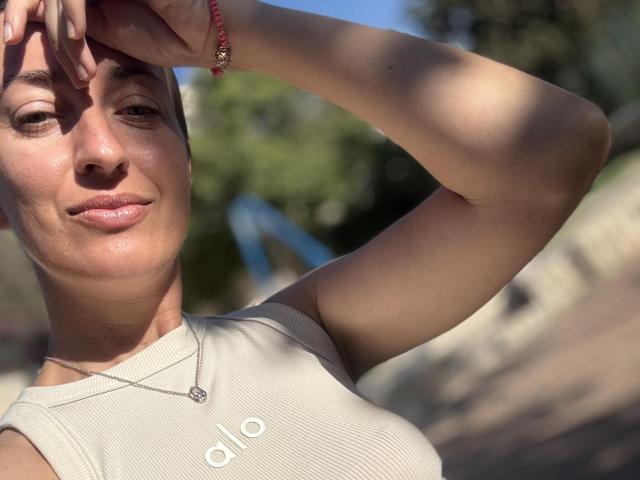 AskaSunshine with short brown hair, fair skin, small tits, wearing a beige ribbed top, sunlit, holding hand up, red bracelet, necklace, slight smile, outdoor light.