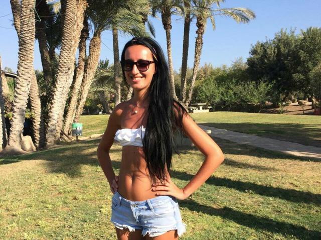 AskaSunshine with long black hair, wearing dark sunglasses, a white strapless top, and blue denim shorts, standing in a sunny park with palm trees and green grass.