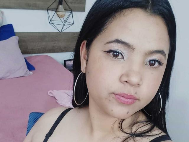 EmilyXGrace with black hair, brown eyes, and hoop earrings, wearing a black top, has puffy lips and a small nose piercing, with subtle skin blemishes, in a selfie.