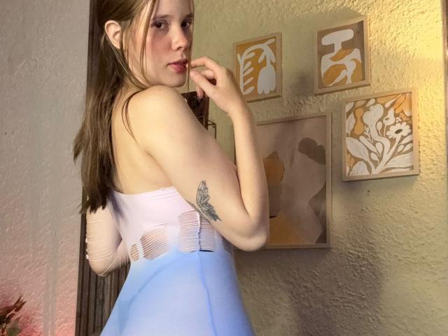 ArielLoveX with pale skin, brown hair, and a small tattoo on her upper arm, wearing a white and blue strapless dress, standing in a room, biting her finger.