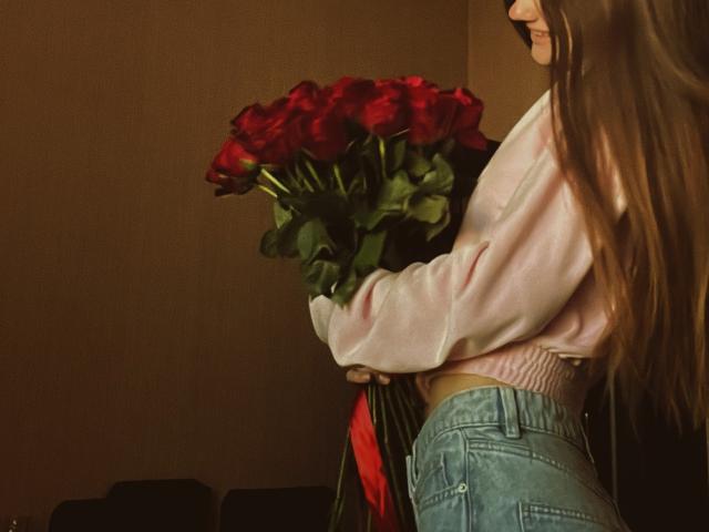 LadyJRouses with long brown hair, pink top, and blue jeans, holding a bouquet of red roses, standing side profile, showing her ass, in a dimly lit room.