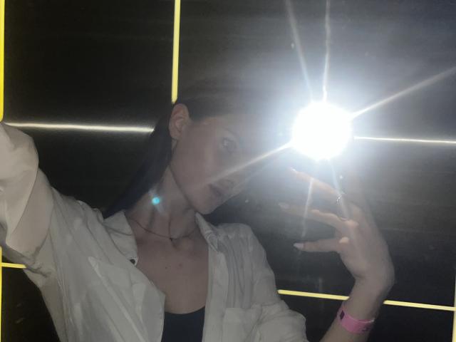 LadyJRouses with dark hair, wearing a white shirt, black top, and pink wristband, taking a selfie with a bright flash, blue earring, and dark lipstick, looking at camera.
