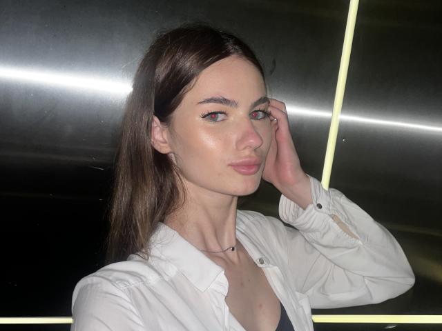 LadyJRouses with shiny skin, brown hair, and pink eyeshadow, wearing a white shirt, hand in hair, looking at camera, slight smile, neon light in background.