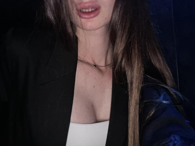 LadyJRouses with light skin and brown hair, wearing a black jacket and white top, showing cleavage and a delicate necklace, with slightly parted pink lips.