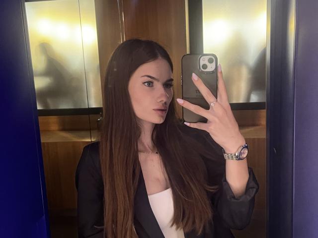 LadyJRouses with long brown hair, wearing a black blazer over a white top, taking a mirror selfie with a gold phone, showing a ring and watch. Boobs visible.