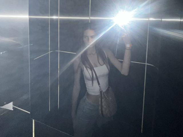 LadyJRouses with long brown hair, small tits, wearing a white crop top and blue jeans, holding a camera with a bright flash, standing in a dark tiled room.