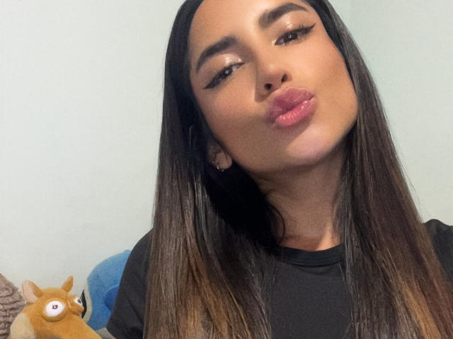 SofyaLane with long brown hair, pouty pink lips, and winged eyeliner, kissing the camera. She's wearing a black shirt, and a cute stuffed fox is on the left.
