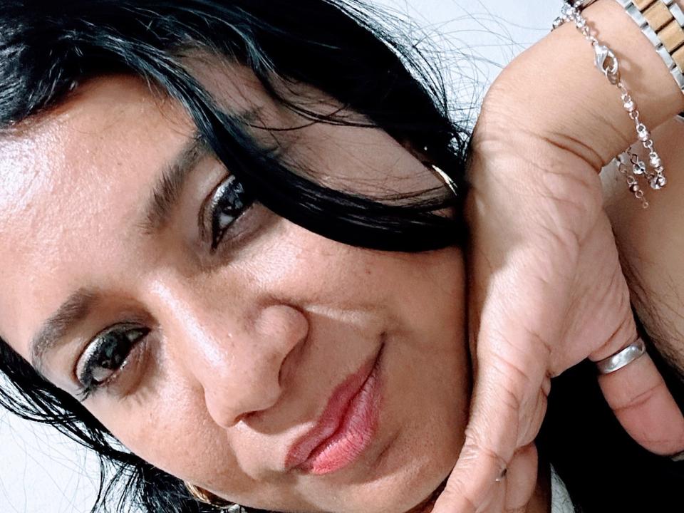 TrishQuinn's face with dark, wavy hair, brown eyes, and a slight smile. She's resting her chin on her hand, wearing a silver ring and a bracelet with small charms.