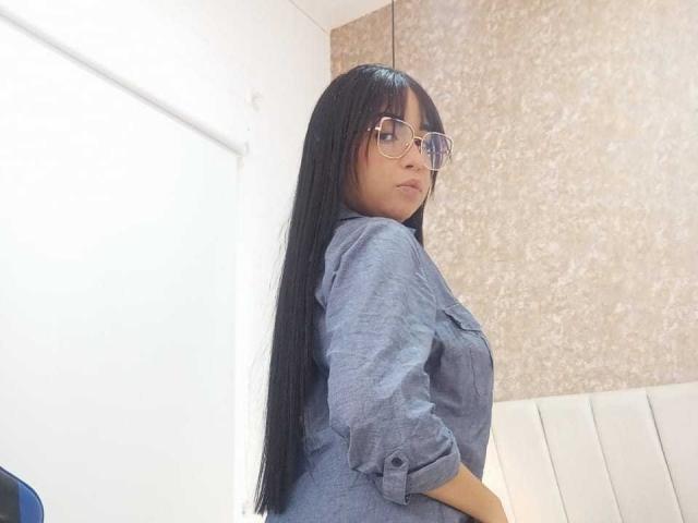 AliceSmity with long black hair, glasses, and a blue button-up shirt, standing with her back to the camera, showing her ass. She's looking over her shoulder.