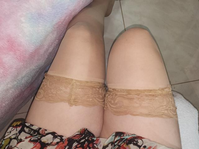 Las muslos de Akkanee, cubiertos con medias de encaje color beige, su trasero ligeramente visible en un vestido floral de colores, sentada en un suelo de baldosas con una manta rosa y blanca..