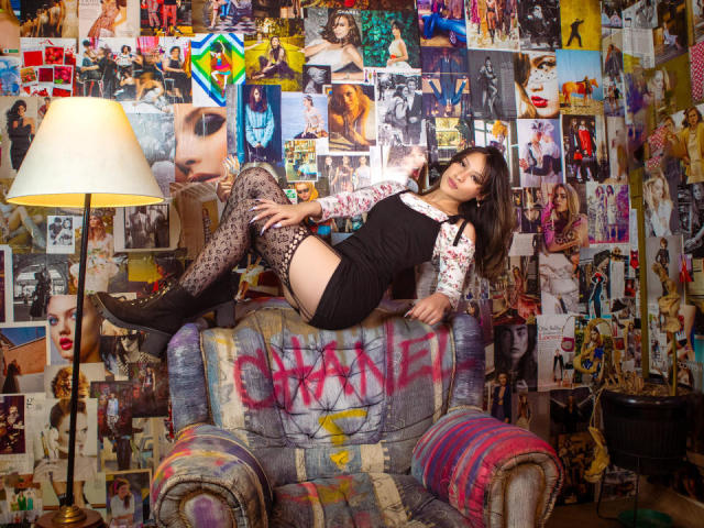 GabriellesRusso lounges on a graffiti-covered armchair, wearing a black dress, white floral shirt, and leopard print thigh-highs, with her ass raised and legs crossed, under a lamp.