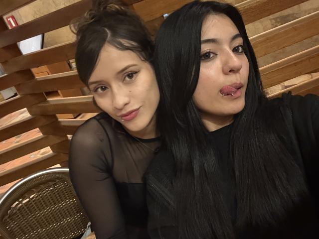 GabriellesRusso with brown hair in a bun, black sheer top, and GabriellesRusso with black hair, black top, and puckered lips, sitting on wooden bench.