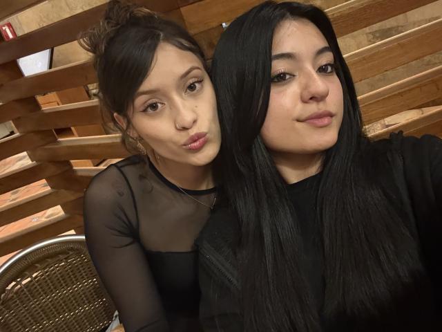 GabriellesRusso with pouty lips, black sheer top, and brown hair, leaning on GabriellesRusso with long black hair, black shirt, and slightly smiling. Wooden slats in background.