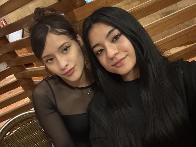 GabriellesRusso with black hair and GabriellesRusso with brown hair, both with small tits, wearing black sheer and black shirts, smiling, wooden slats in background, selfie.