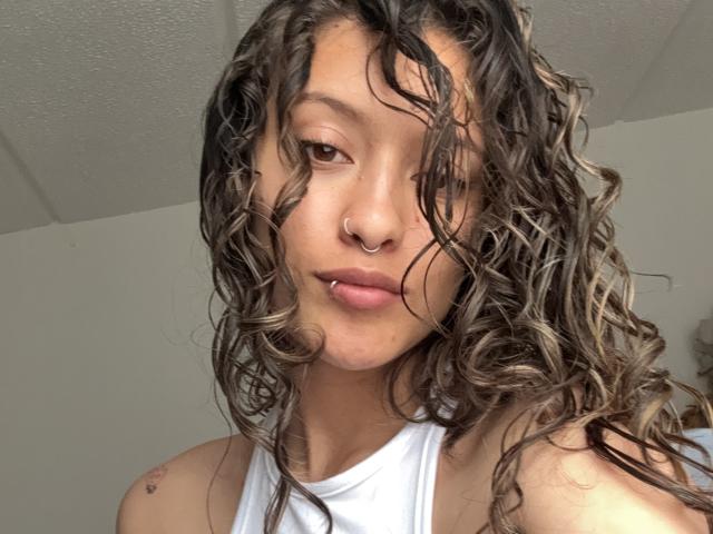 GabriellesRusso with curly brown hair, nose ring, and lip piercing, wearing a white tank top, looking at the camera with a slightly pouty expression.