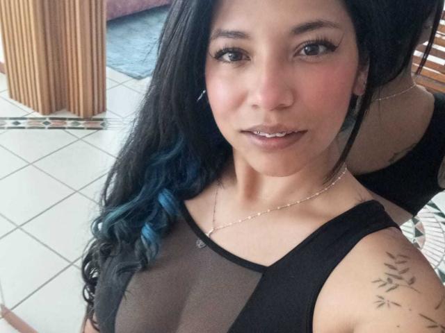PaulinaColemann has black wavy hair with blue tips, wearing a sheer black top, showing her tits, small tattoos on her shoulder, and a silver necklace. She's looking at the camera.