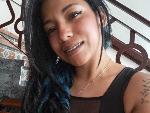 PaulinaColemann has black hair with blue tips, a nose ring, and a silver necklace. She's smiling with metallic teeth, wearing a black top, and showing a tattoo on her arm.