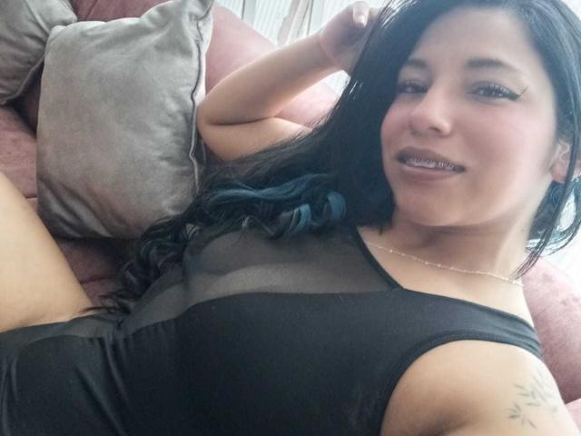 PaulinaColemann with black dress, sheer chest, blue-tipped hair, lying on couch, arm raised, smiling, showing teeth, small tattoo on right arm, pink couch, gray pillow.