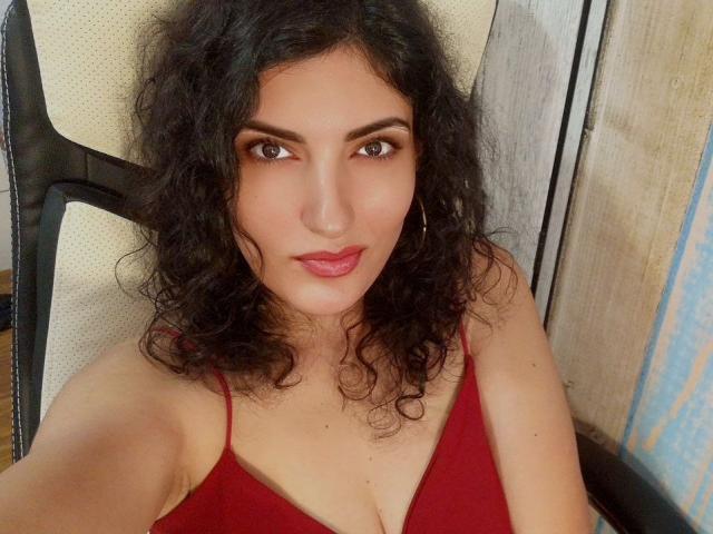 AddictedToUTS has curly black hair, brown eyes, and is wearing a red tank top with visible cleavage. She's sitting in a chair with a neutral expression.