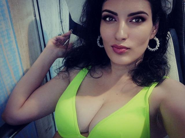 AddictedToUTS has large, shiny tits in a neon green bikini top, wearing big hoop earrings, with dark curly hair and pink lipstick, looking at the camera.