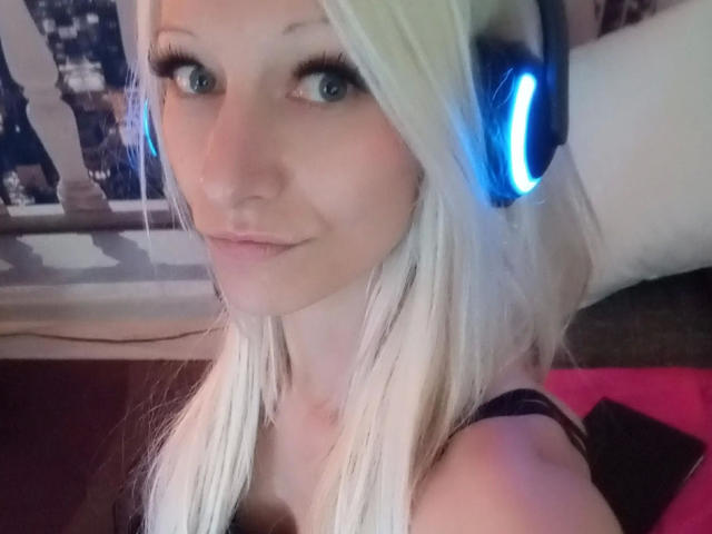 Kitty-Deluxe-hot with blonde hair, blue headphones, and a cute smile, showing her tits in a selfie, with long eyelashes and a subtle pink lip.