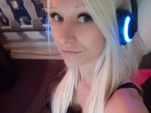 Kitty-Deluxe-hot with blonde hair, blue headphones, and a slight smile, showing her tits in a black top. She's got green eyes and a cute, innocent face.