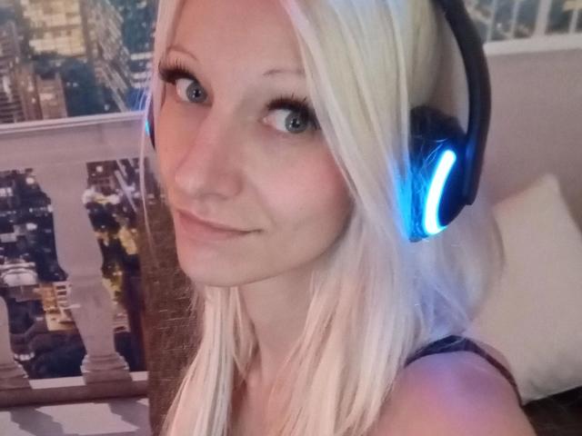 Kitty-Deluxe-hot with blonde hair, blue headphones, and a slight smile, showing her cute face and big green eyes. Her tits are not visible. She's looking at the camera.