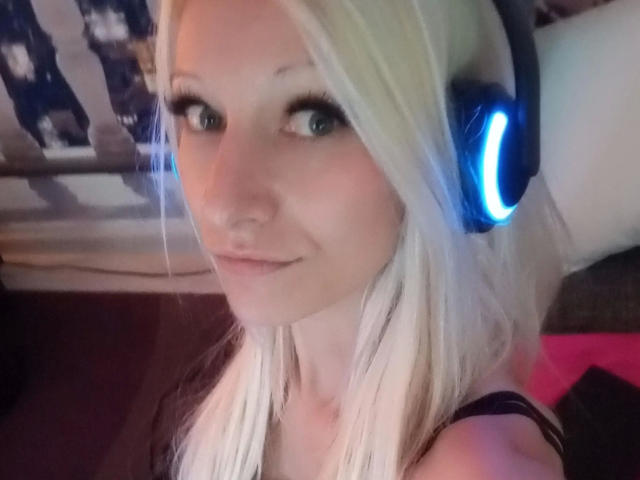 Kitty-Deluxe-hot with blonde hair, blue headphones, and a slight smile, showing her cute face and small nose. She has green eyes and a subtle makeup look.