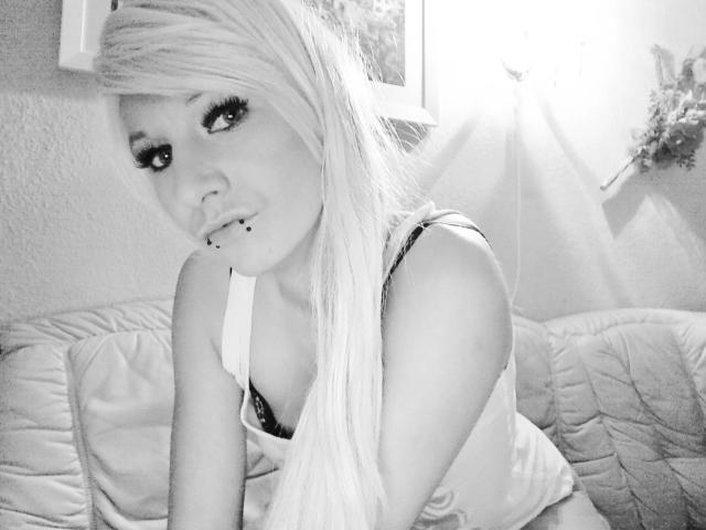 Kitty-Deluxe-hot with long blonde hair, heavy eyelashes, and a nose piercing, wearing a white top, showing her small tits and black bra, sitting on a quilted couch.