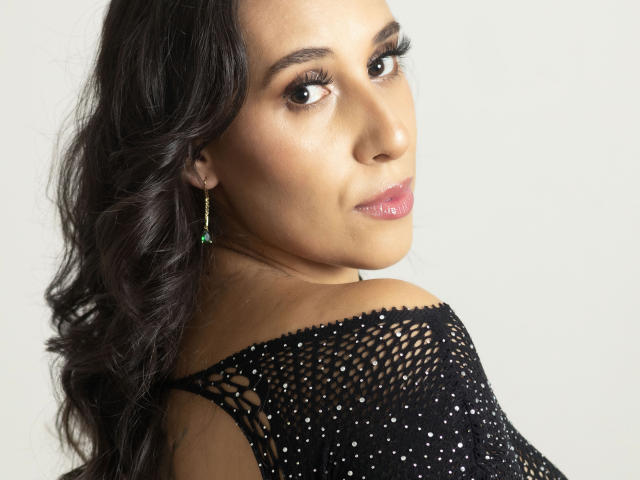 BellaBrownX with dark wavy hair, brown eyes, and pink lipstick, wearing a black, sparkly, off-shoulder top, green earrings with a small gem, looking over her shoulder.