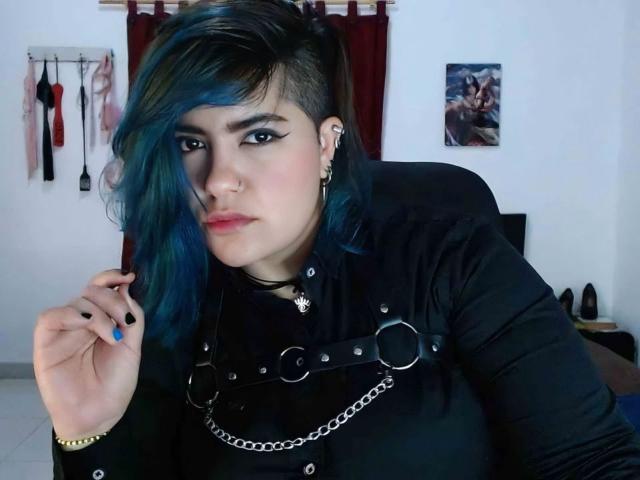 ZussanGravereign has blue and black hair, black eyeliner, black nails, black top with metal rings, and chain, sitting, looking at camera, wearing nose ring, and black choker.