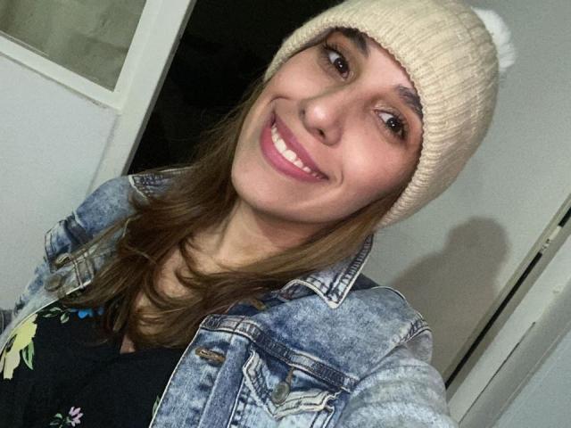 BellaBrownX with a smile, wearing a beige knit beanie, blue denim jacket, and black top, showing her tits slightly in a casual selfie.