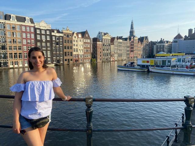 BellaBrownX with off-shoulder blue top, small tits, denim shorts, standing by canal, smiling, brown hair, casual, sunny, boats, historic buildings in background, metal railing in foreground.
