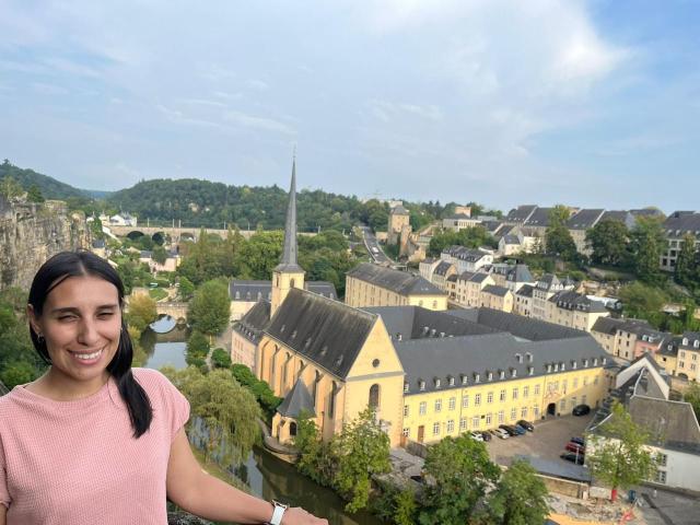 BellaBrownX with dark pigtails, pink sweater, smiling, standing in front of a yellow historic building with a church steeple, green trees, and a small town under a blue sky.